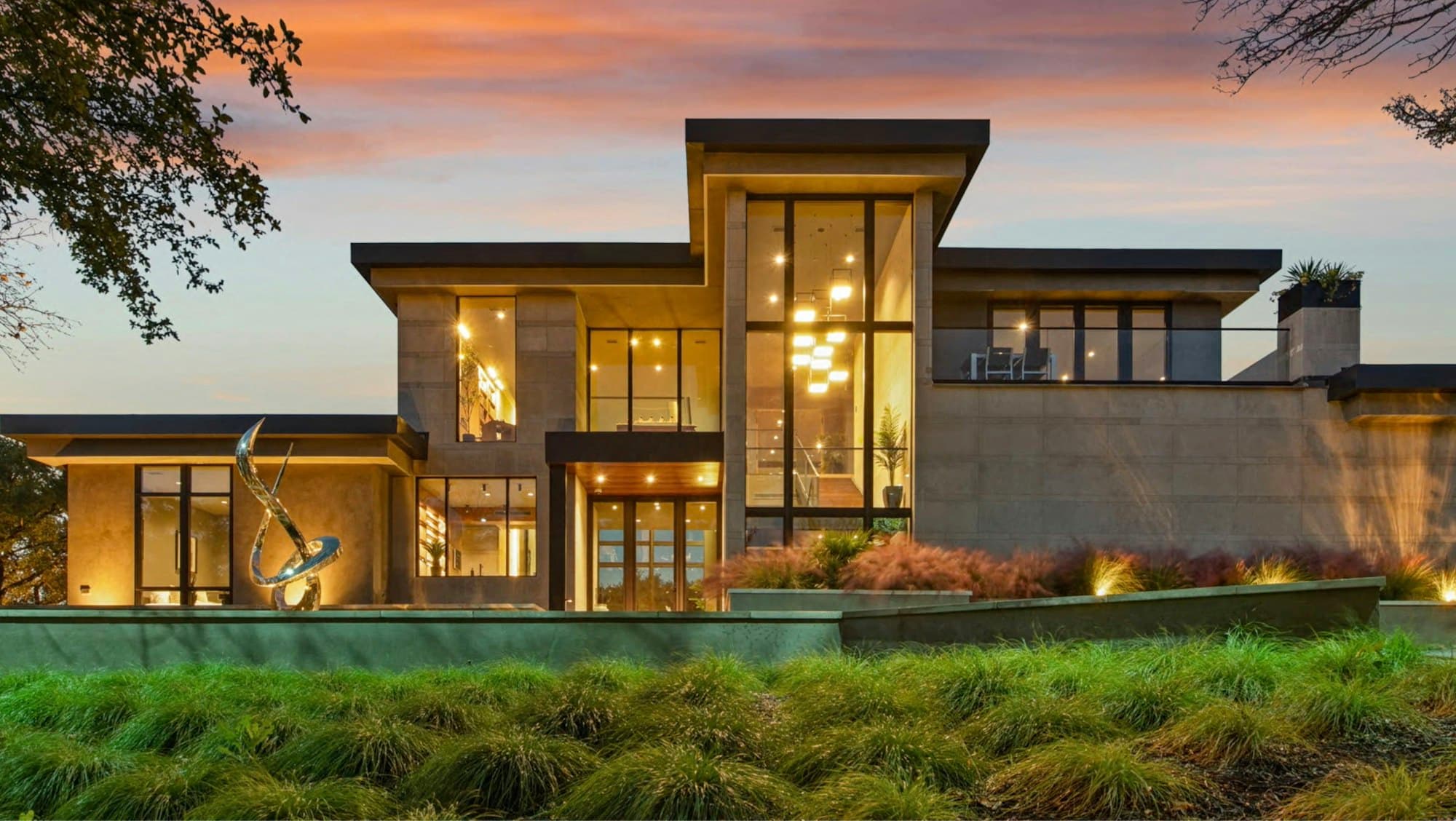 A low-slung contemporary residence at golden hour with warm interior lighting and layered landscape planting