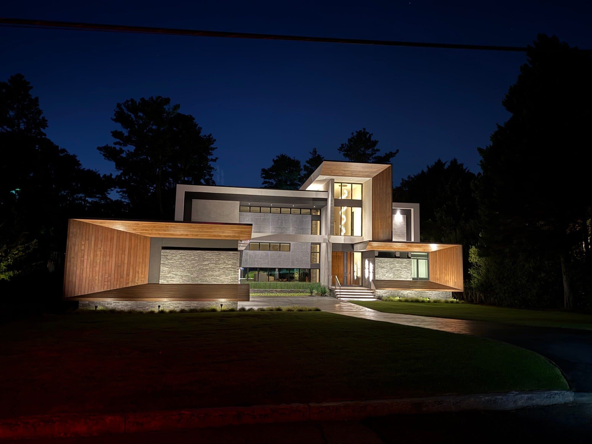 A modern home at night with warm facade lighting and a floodlit lawn