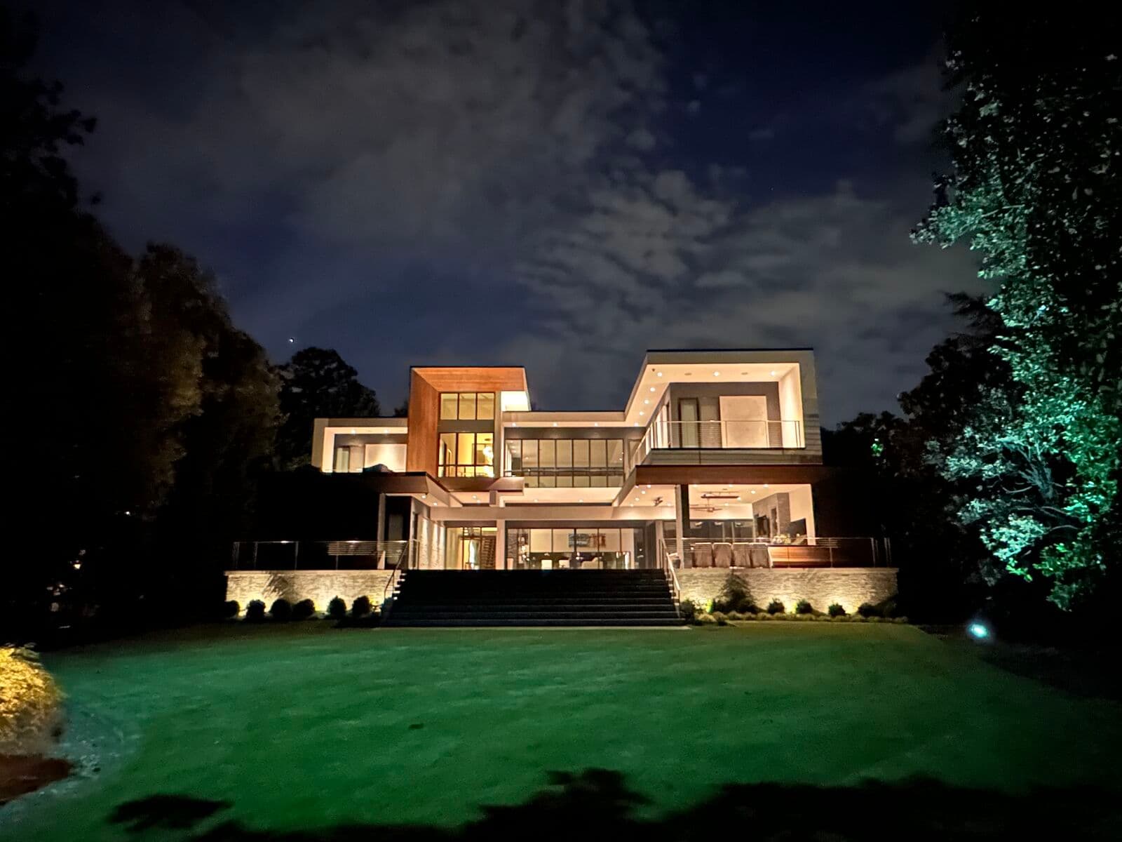 A modern home at night with a warmly lit facade, uplit trees, and a floodlit rear lawn